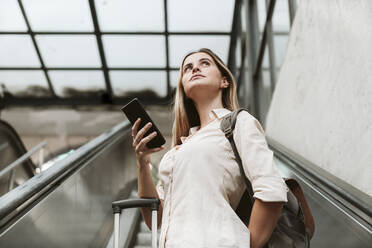 Young woman wearing backpack looking away standing on escalator at railroad station - EBBF04168