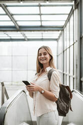 Happy young woman wearing backpack standing by escalator at railroad station - EBBF04169