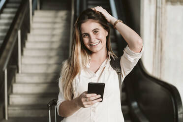 Glückliche junge Frau hält ihr Smartphone in der Hand, während sie mit der Hand im Haar steht - EBBF04170