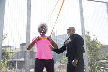 Man helping woman exercising with resistance band at park on sunny day - PMF01965