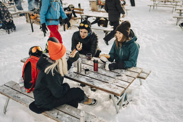 Männliche und weibliche Freunde unterhalten sich beim Essen eines Hotdogs in einem Skigebiet im Winter - MASF24320