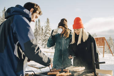 Man preparing sausages while female friends standing at ski resort - MASF24323