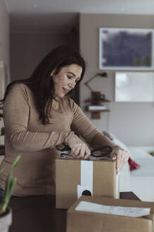 Mittlere erwachsene Frau beim Auspacken eines Kartons mit einer Schere zu Hause - MASF24389