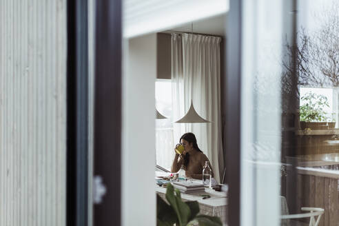 Weibliche Fachkraft trinkt Kaffee durch ein Fenster im Büro zu Hause gesehen - MASF24400