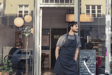 Männlicher Besitzer schaut weg, während er mit den Händen in den Taschen am Eingang eines Cafés steht - MASF24474