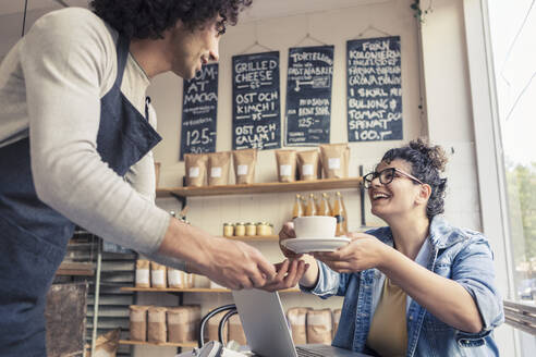 Männlicher Kellner serviert einer fröhlichen Kundin in einem Café Kaffee - MASF24475