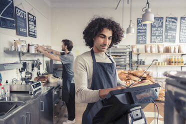 Männlicher Besitzer benutzt die Registrierkasse, während ein Mitbesitzer im Café arbeitet - MASF24478