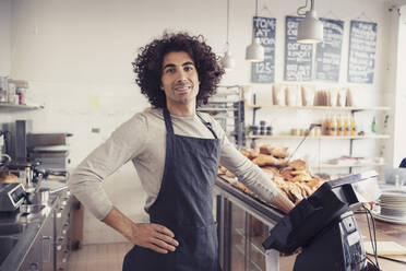 Porträt eines männlichen Unternehmers, der mit der Hand an der Hüfte an der Kasse eines Cafés steht - MASF24485