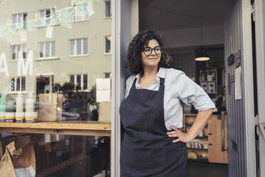 Glückliche Unternehmerin, die mit der Hand auf der Hüfte steht und den Blick vom Ladeneingang abwendet - MASF24497