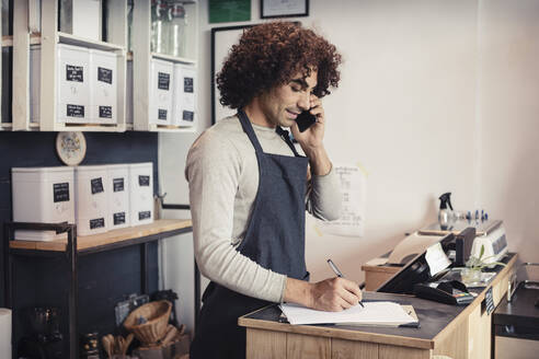 Verkäufer, der mit seinem Handy telefoniert, während er an der Kasse schreibt - MASF24525