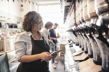 Female owner checking inventories with digital tablet while male salesman assisting customer in shopping at organic stor - MASF24530