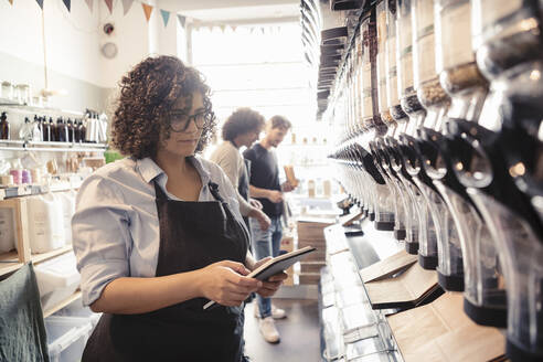 Eine Frau prüft die Bestände mit einem digitalen Tablet, während ein männlicher Verkäufer den Kunden beim Einkauf im Bioladen hilft - MASF24530
