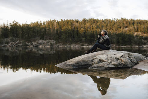 Mature female explorer contemplating while sitting on rock by lake and trees - MASF24549