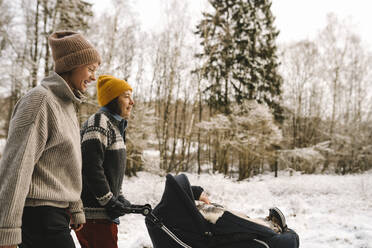Mature woman pushing baby stroller while walking with girlfriend during winter - MASF24721