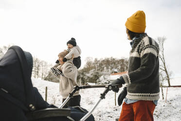 Smiling woman carrying daughter on shoulder while girlfriend walking with carriage during winter - MASF24722