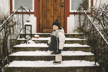 Mädchen in voller Länge, das warme Kleidung trägt, während es im Winter auf einer schneebedeckten Treppe steht - MASF24726