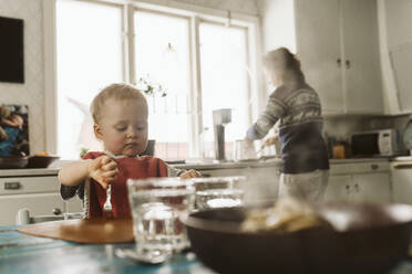 Mädchen sitzt am Esstisch, während die Mutter in der Küche Essen zubereitet - MASF24731