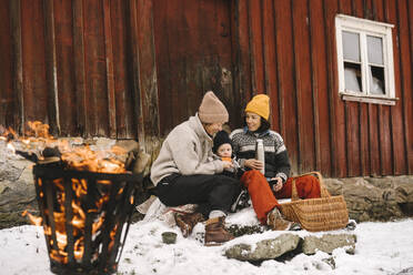 Mature woman sitting with girlfriend feeding daughter against cottage during winter - MASF24744