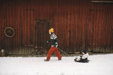 Ältere Frau, die ihre Tochter auf einem Schlitten zieht, während sie im Winter an einer Hütte vorbeiläuft - MASF24771