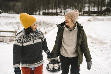 Lächelndes lesbisches Paar, das sich gegenseitig ansieht, während es seine Tochter durch den Schnee zieht - MASF24778