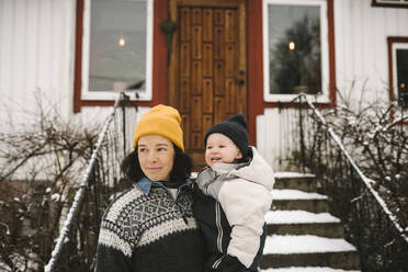 Lächelnde Mutter, die ihre Tochter vor einem Haus trägt, während sie im Winter wegschaut - MASF24782