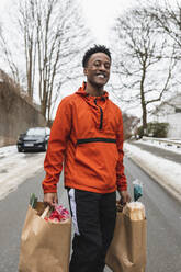 Portrait of smiling essential service male carrying paper bags while walking on road - MASF24817