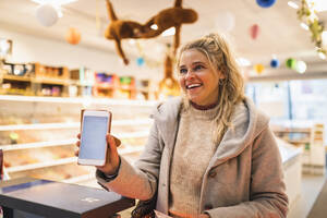 Glückliche blondhaarige Frau zeigt Smartphone im Geschäft - MASF24838