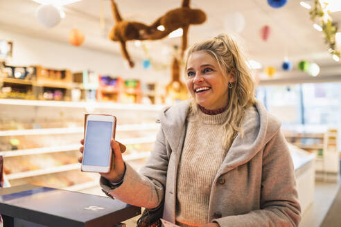 Happy blond hair woman showing smart phone at store - MASF24838