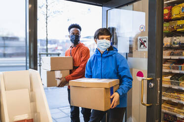 Male and female delivery colleagues carrying cardboard boxes while entering retail store during pandemic - MASF24842