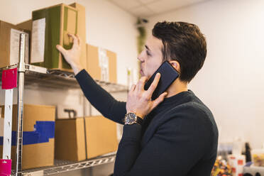 Male entrepreneur keeping box on shelf while talking through smart phone in store - MASF24843