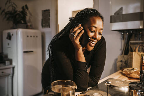 Junge Frau spricht über ihr Smartphone, während sie sich auf den Küchentisch stützt - MASF24878