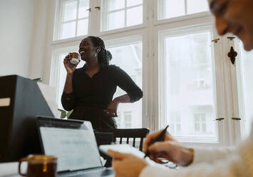 Lächelnde Geschäftsfrau mit Kaffeetasse am Fenster stehend, während ein männlicher Kollege im Heimbüro schreibt - MASF24883