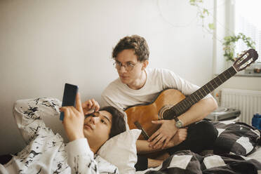Mann mit Gitarre guckt, während männlicher Freund Smartphone im Schlafzimmer benutzt - MASF24953
