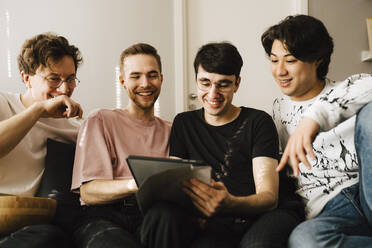 Lächelnder Mann, der ein digitales Tablet benutzt, während er sich mit männlichen Freunden im Wohnzimmer austauscht - MASF24961