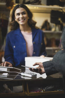 Eine lächelnde Inhaberin betrachtet einen Kunden, der in einem Geschäft kontaktlos bezahlt - MASF24978