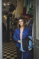 Lächelnde junge Besitzerin, die Kleidung in der Hand hält, während sie an der Tür eines Geschäfts steht - MASF25006