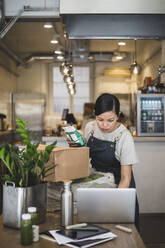 Weibliche Angestellte arbeitet am Laptop und hält ein Lebensmittelpaket in einem Geschäft - MASF25058