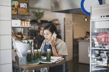 Asiatische Unternehmerin, die über ein Smartphone spricht, während sie in einem Geschäft an einem Laptop arbeitet - MASF25064