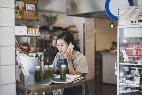 Asiatische Unternehmerin, die über ein Smartphone spricht, während sie in einem Geschäft an einem Laptop arbeitet - MASF25064