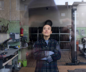 Weiblicher Mechaniker, der mit verschränkten Armen in einer Werkstatt steht und nach oben schaut - KNSF08854