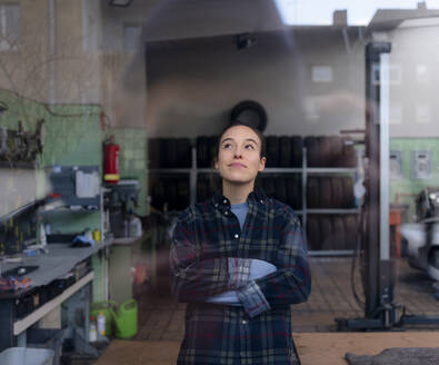 Weiblicher Mechaniker, der mit verschränkten Armen in einer Werkstatt steht und nach oben schaut - KNSF08854