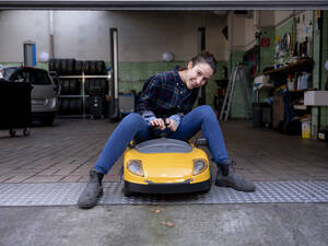 Smiling female mechanic sitting on toy car at car repair shop - KNSF08872