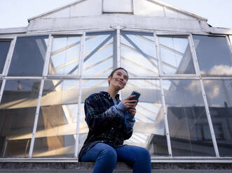 Smiling woman with mobile phone sitting on rooftop - KNSF08875