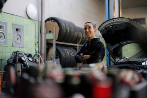 Smiling female mechanic with arms crossed looking away while leaning on car - KNSF08883