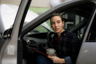 Mechanikerin mit Kaffeetasse, die wegschaut, während sie im Auto sitzt - KNSF08894