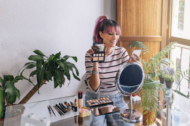 Smiling female influencer showing make-up palette while vlogging through mobile phone at home - EGHF00069
