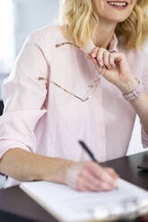 Female professional holding eyeglasses while writing on paper at home office - WPEF04939