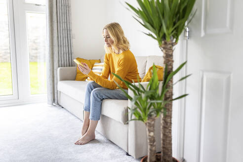 Blonde Frau benutzt ein Smartphone, während sie zu Hause auf dem Sofa sitzt - WPEF04970