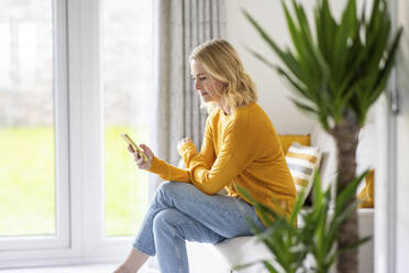 Woman sitting with legs crossed at knee using smart phone on sofa at home - WPEF04971