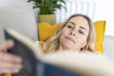 Woman reading book while relaxing on sofa at home - WPEF04982